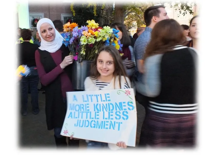 A young volunteer holding a sign reading "A little more kindness, a little less judgment," with flowers, at a Murfreesboro Loves gathering